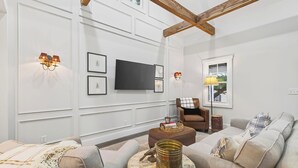 Ein Wohnzimmer mit einem großen Flachbildfernseher, zwei Wandlampen, einem braunen Ledersessel, einem beigen Sofa und einem runden Couchtisch mit einer Dekoration.