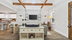 Ein großzügiges Wohnzimmer mit hohen Decken, Holzbalken, einem Flachbildfernseher, einem weißen Konsolentisch und einer gemütlichen Sitzecke.