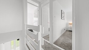 Un couloir blanc avec une rambarde en bois, un tableau encadré au mur et une lampe sur une table de chevet.
