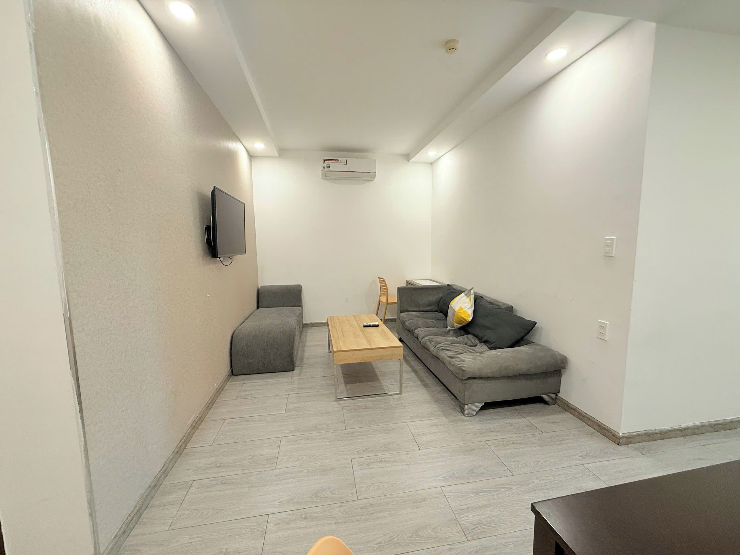 A compact living space with a sofa, a small table, a TV mounted on the wall, and a ceiling-mounted air conditioner.