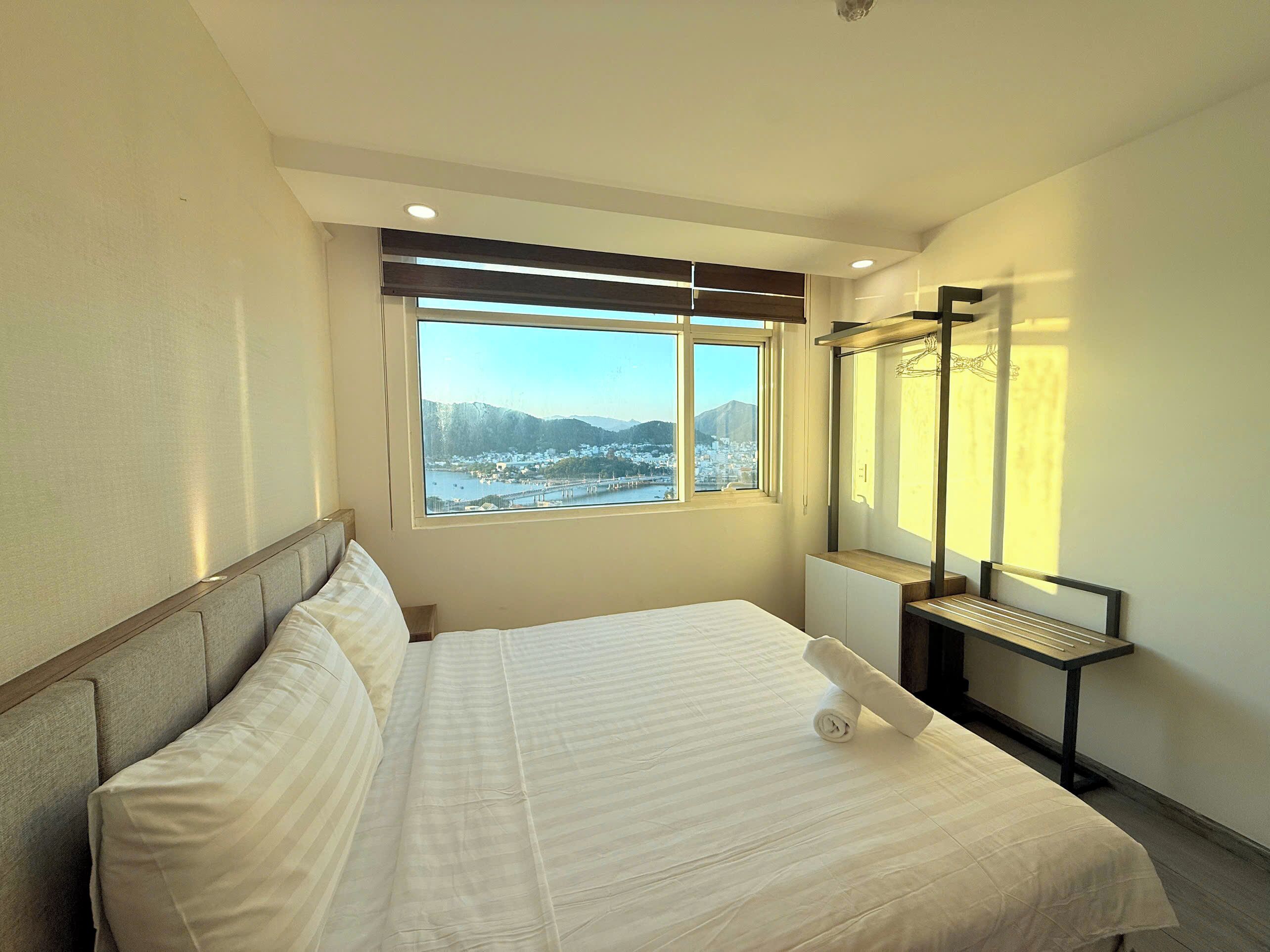 Una habitación de hotel con una cama, una ventana con vistas a un pueblo y montañas, un espejo y una pequeña mesa.