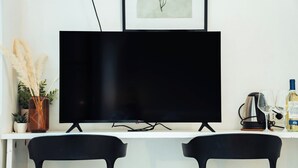 Un televisor de pantalla plana sobre un soporte blanco, dos sillas negras, un estante blanco con una planta, un cuadro enmarcado y una tetera a la derecha.