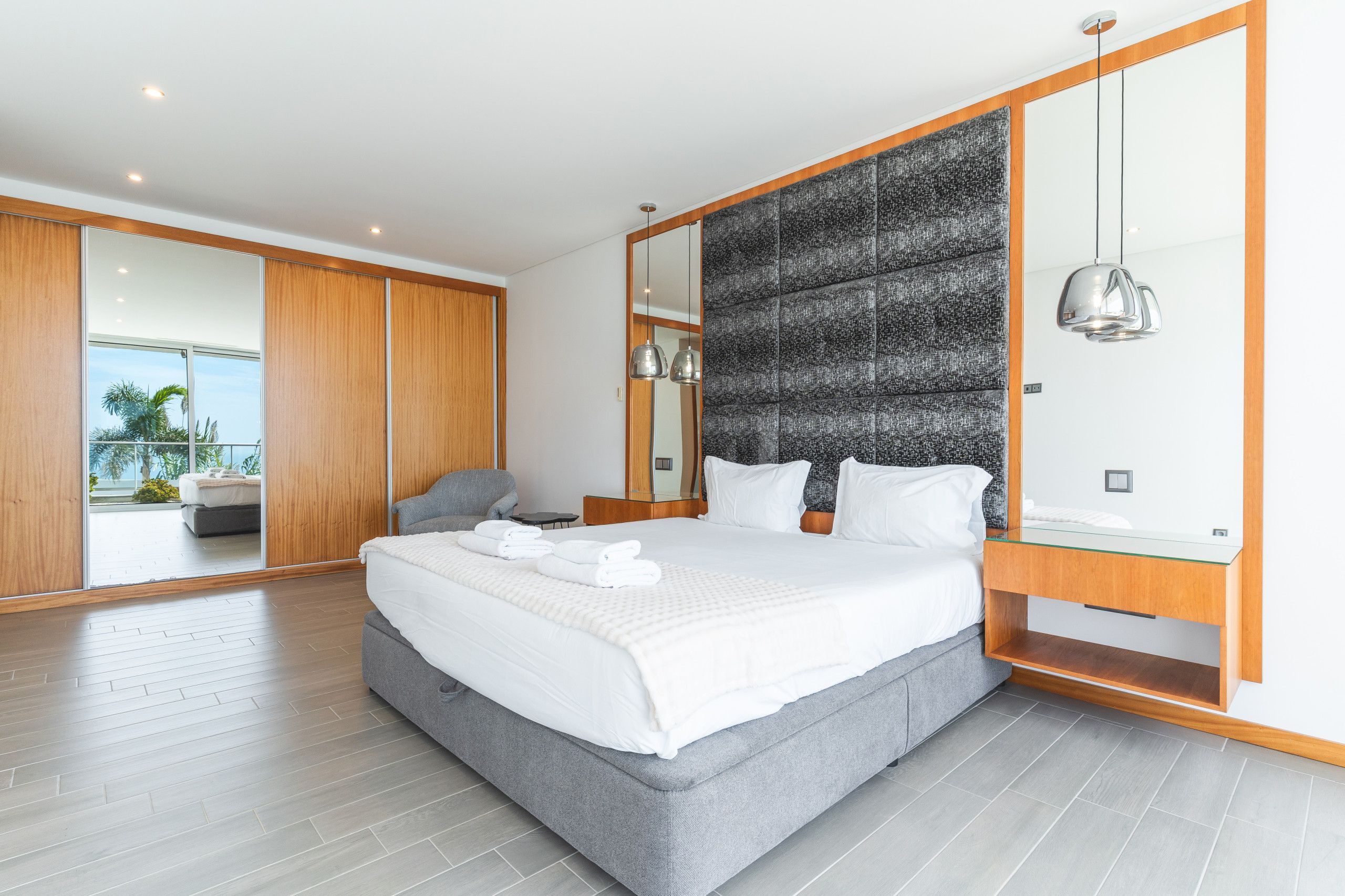 Een moderne slaapkamer met een groot bed, een grijze bank, een houten garderobe en een grote spiegel.