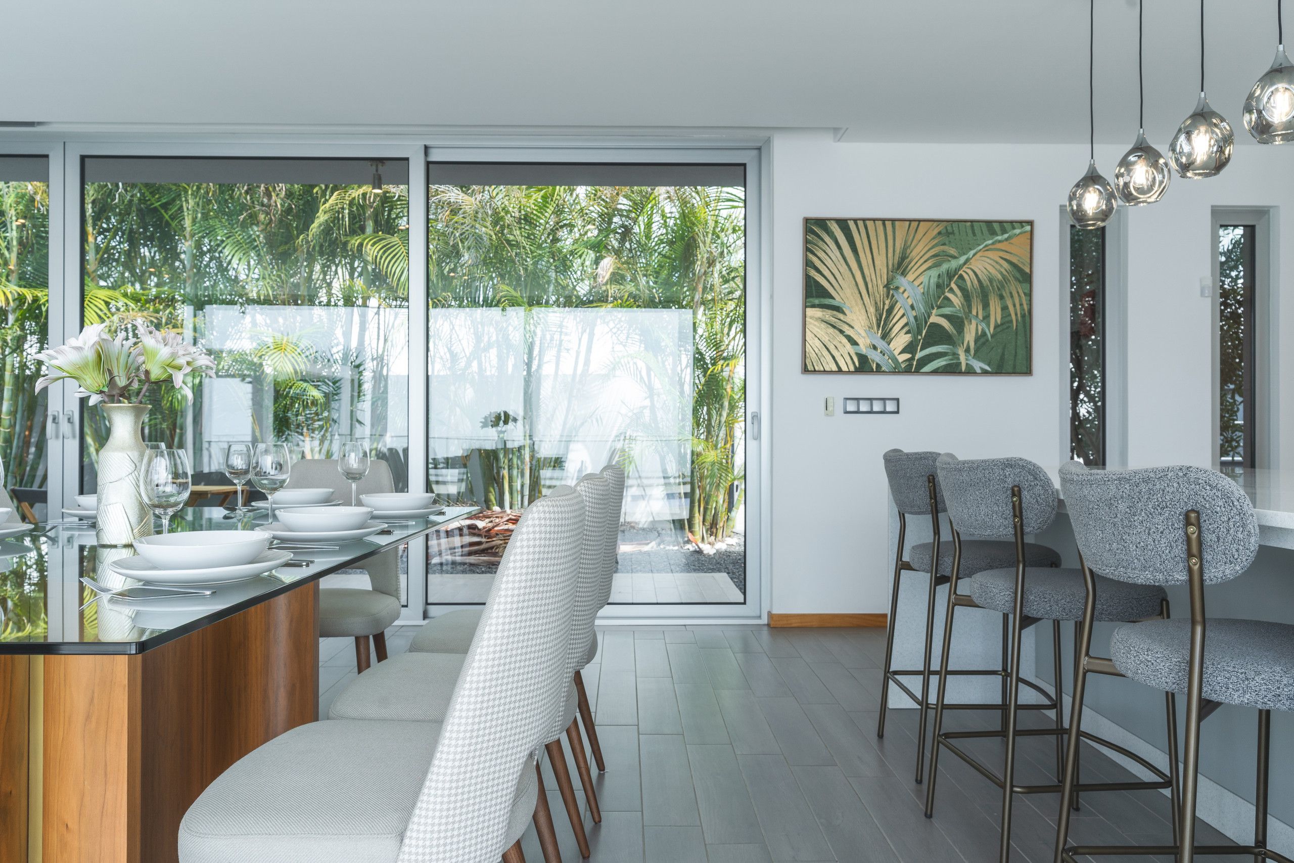 A modern dining area with a glass table, white chairs, and a view of greenery outside.