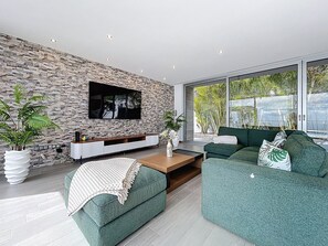 A modern living room with a stone wall, a flat-screen TV, green sofas, and large windows with a view of greenery. - Villa Vista Atlântica by Atlantic Holiday (Ribeira Brava)