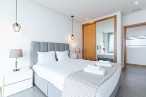 Une chambre d’hôtel moderne avec un grand lit, des lampes de chevet, une armoire en bois et une salle de bain visible à travers une porte ouverte.