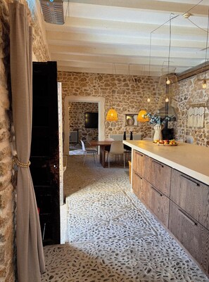 Une cuisine aux murs de pierre, au décor rustique, avec un plan de travail en bois, un sol en pierre et des luminaires suspendus.