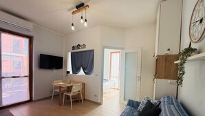 A room with a wooden floor, a sofa, a TV, a table with chairs, a window with curtains, and a door leading to another room. - Terrazza vicino alla spiaggia in centro (Marina di Andora)