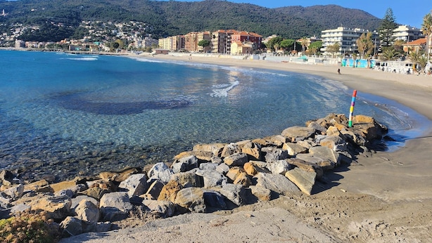 A coastal beach with clear water, rocky shoreline, and buildings in the background. - Terrazza vicino alla spiaggia in centro (Marina di Andora)
