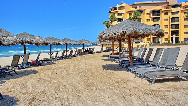 A beach with rows of sun loungers and thatched umbrellas, a clear blue sky, and a hotel building in the background. - PrincesaB108 GroundFlr Grill ExperienceRockyPoint Oversized Patio (Puerto Penasco)