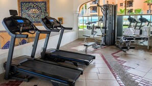 A gym with treadmills, weight machines, and a large window with a view of buildings and palm trees. - PrincesaB108 GroundFlr Grill ExperienceRockyPoint Oversized Patio (Puerto Penasco)
