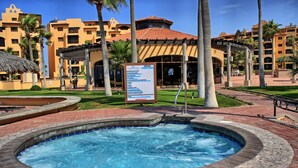 A hot tub surrounded by a brick patio, palm trees, and a building with a tiled roof. - PrincesaB108 GroundFlr Grill ExperienceRockyPoint Oversized Patio (Puerto Penasco)