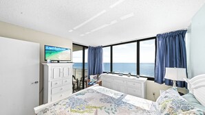 Quarto com uma cama grande, cômoda, televisão e vista para o oceano.