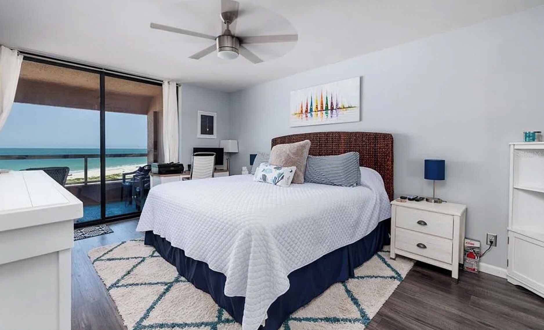Ein Schlafzimmer mit einem Bett, einem Nachttisch, einem Schreibtisch, einem Stuhl, einem Deckenventilator, einem Gemälde und Blick aufs Meer.