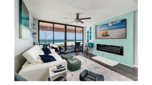 Ein modernes Wohnzimmer mit großem Fenster mit Blick auf den Strand, einem weißen Sofa, einem grünen Hocker und einem flachbildfernseher an der Wand.