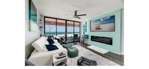 A modern living room with a large window overlooking the beach, a white sofa, a green ottoman, and a flat-screen TV mounted on the wall. - 2-bedroom beach condo, water views in beautiful Sand Key, 4 mins to Clearwater (Clearwater)