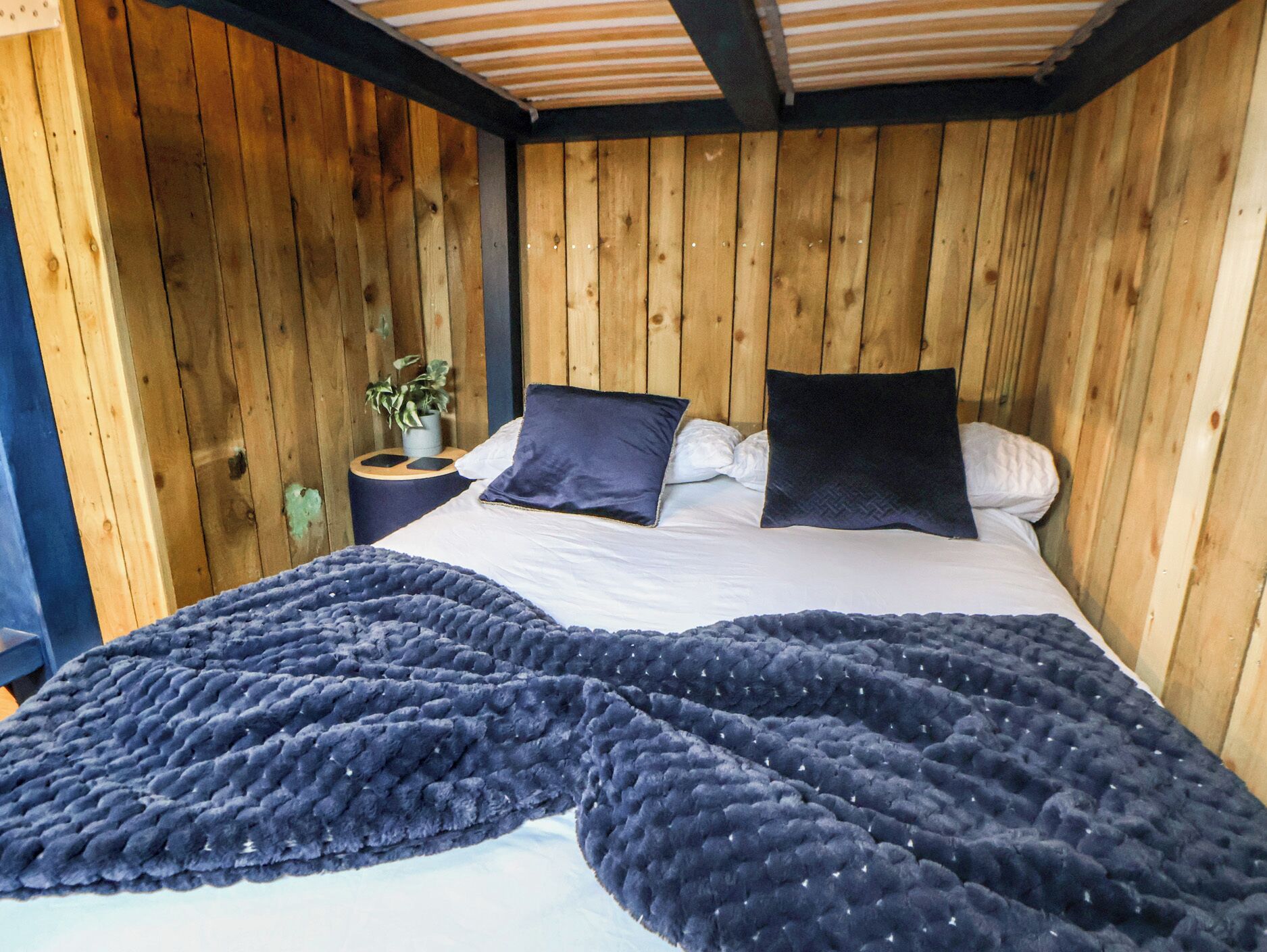 Habitación con paneles de madera, una cama, almohadas, una manta y una mesita con una planta.