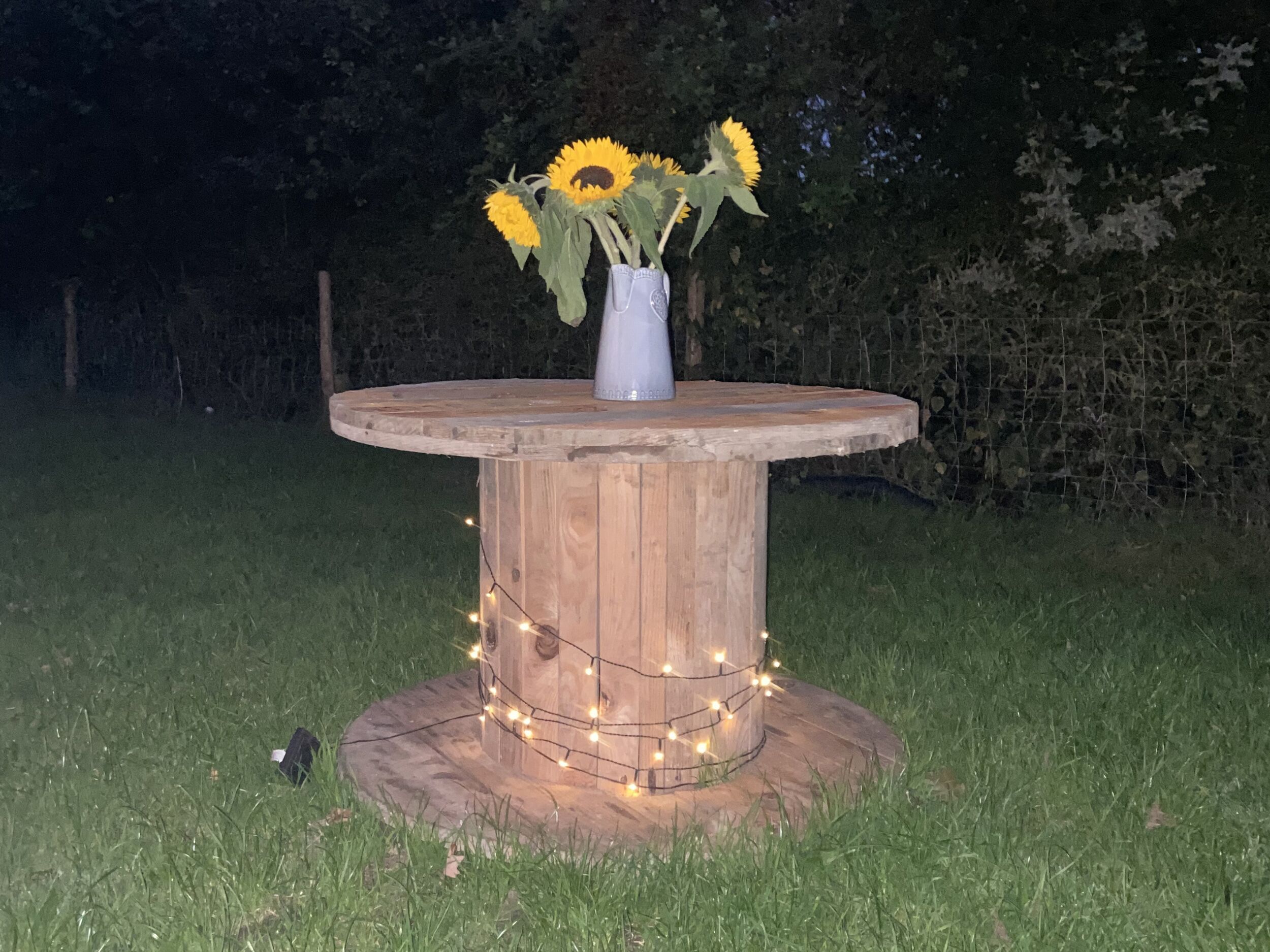 A wooden table with a vase of sunflowers and fairy lights.