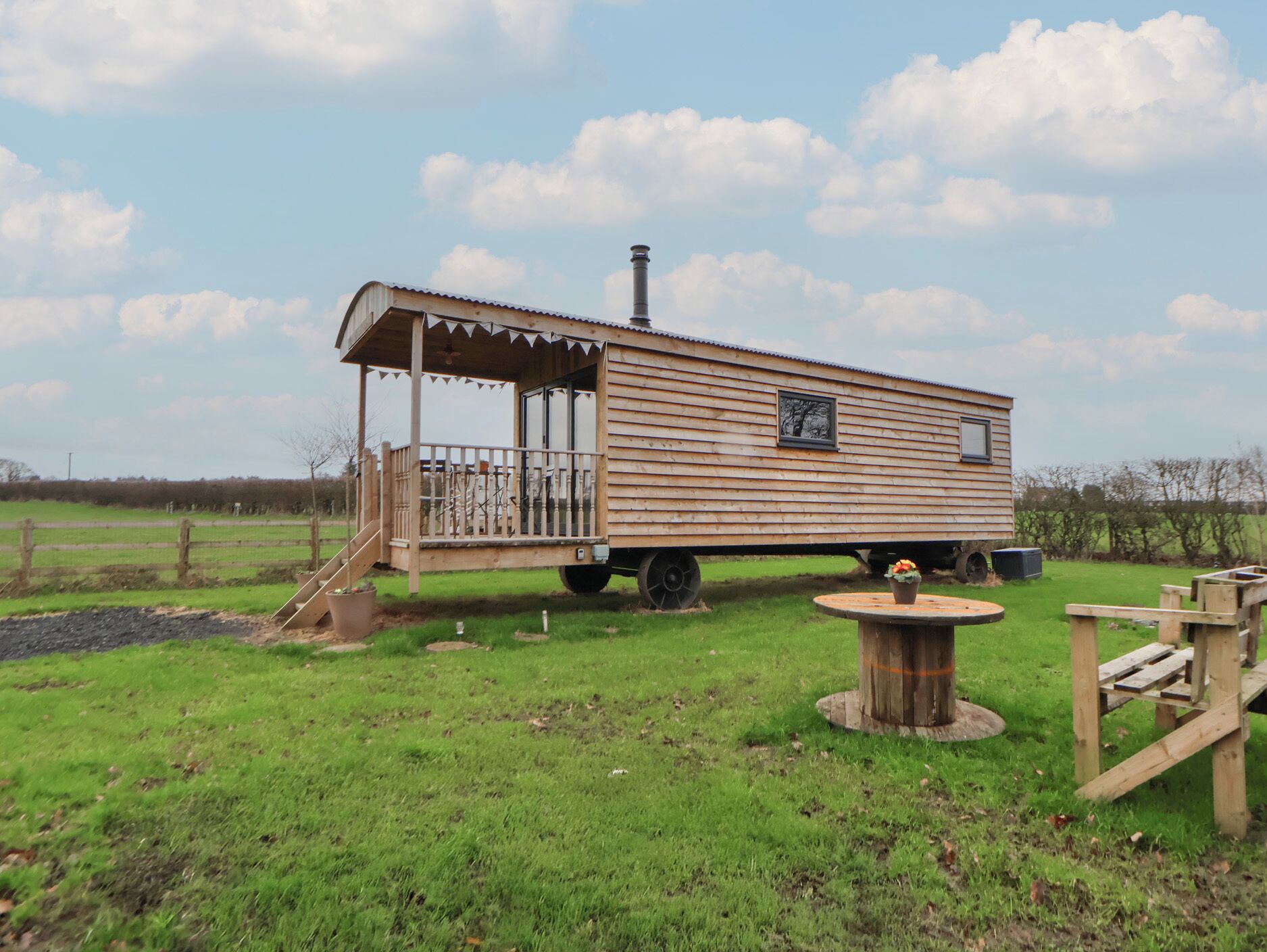 A wooden houseboat with a covered deck and a chimney, situated on a grassy field with a fence in the background.