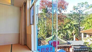 A traditional Japanese-style room with tatami flooring, a shoji screen, and a view of a traditional garden with a thatched-roof structure.