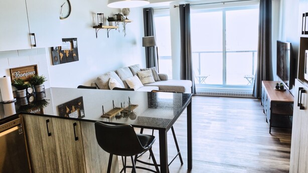 A modern apartment with a kitchen, dining area, and living room. It features a large window, a sofa, a coffee table, and a TV. - Le1006 - Condo moderne (Québec)