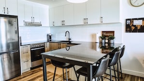 A modern kitchen with white cabinets, a stainless steel refrigerator, a built-in oven, a black countertop, and a bar stool area. - Le1006 - Condo moderne (Québec)