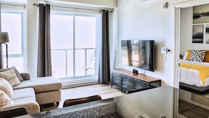 A modern living room with a sofa, a coffee table, a TV, and a balcony with curtains. - Le1006 - Condo moderne (Québec)