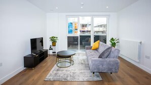 A modern living room with a grey sofa, a black entertainment unit, a small round table, and a large window with a view of a building. - 2 Bed Flat - Sleeps 6 - Parking - Wifi (London)
