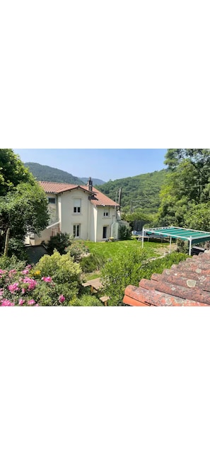 A two-story house with a red-tiled roof, surrounded by lush greenery and a garden with pink flowers. - The blue house (Courniou)