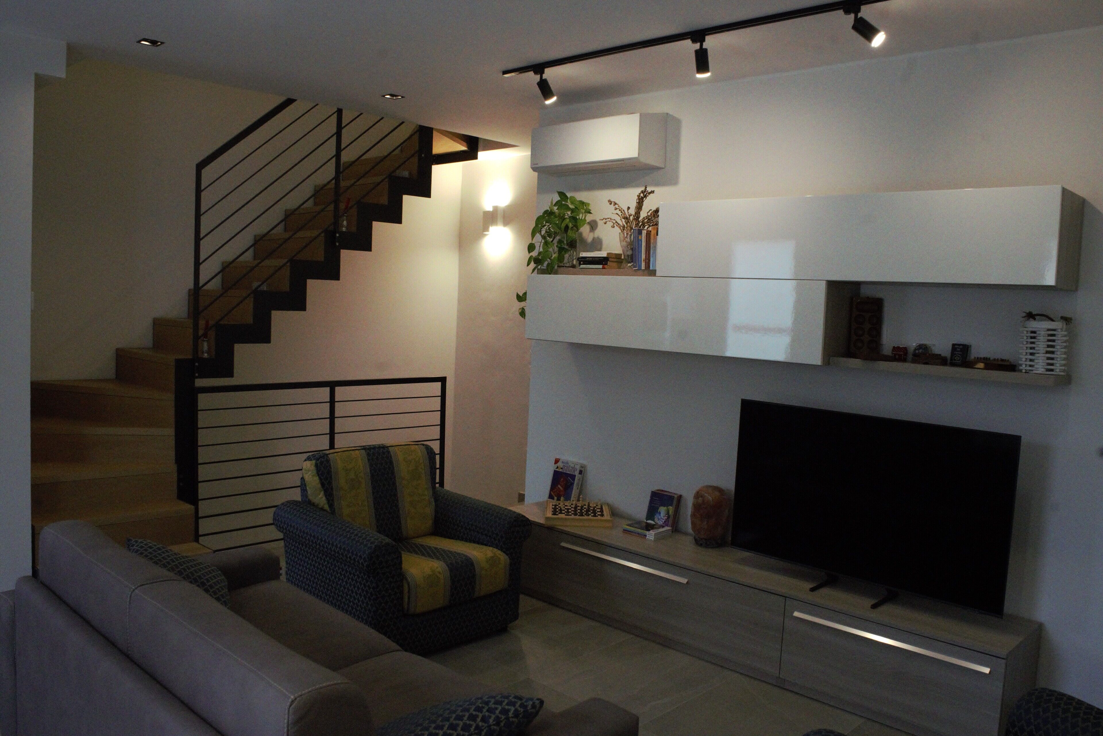 A modern living room with a sofa, a chair, a flat-screen TV, a staircase, and built-in shelving.