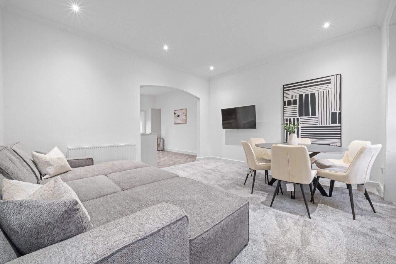 A modern living room with a grey sectional sofa, a dining area with a round table and chairs, a flat-screen TV mounted on the wall, and a large abstract artwork.