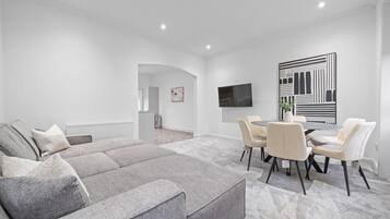 A modern living room with a grey sectional sofa, a dining area with a round table and chairs, a flat-screen TV mounted on the wall, and a large abstract artwork.