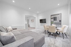 A modern living room with a grey sectional sofa, a dining area with a round table and chairs, a flat-screen TV mounted on the wall, and a large abstract artwork. - Loch Lomond Hillside Retreat (Alexandria)