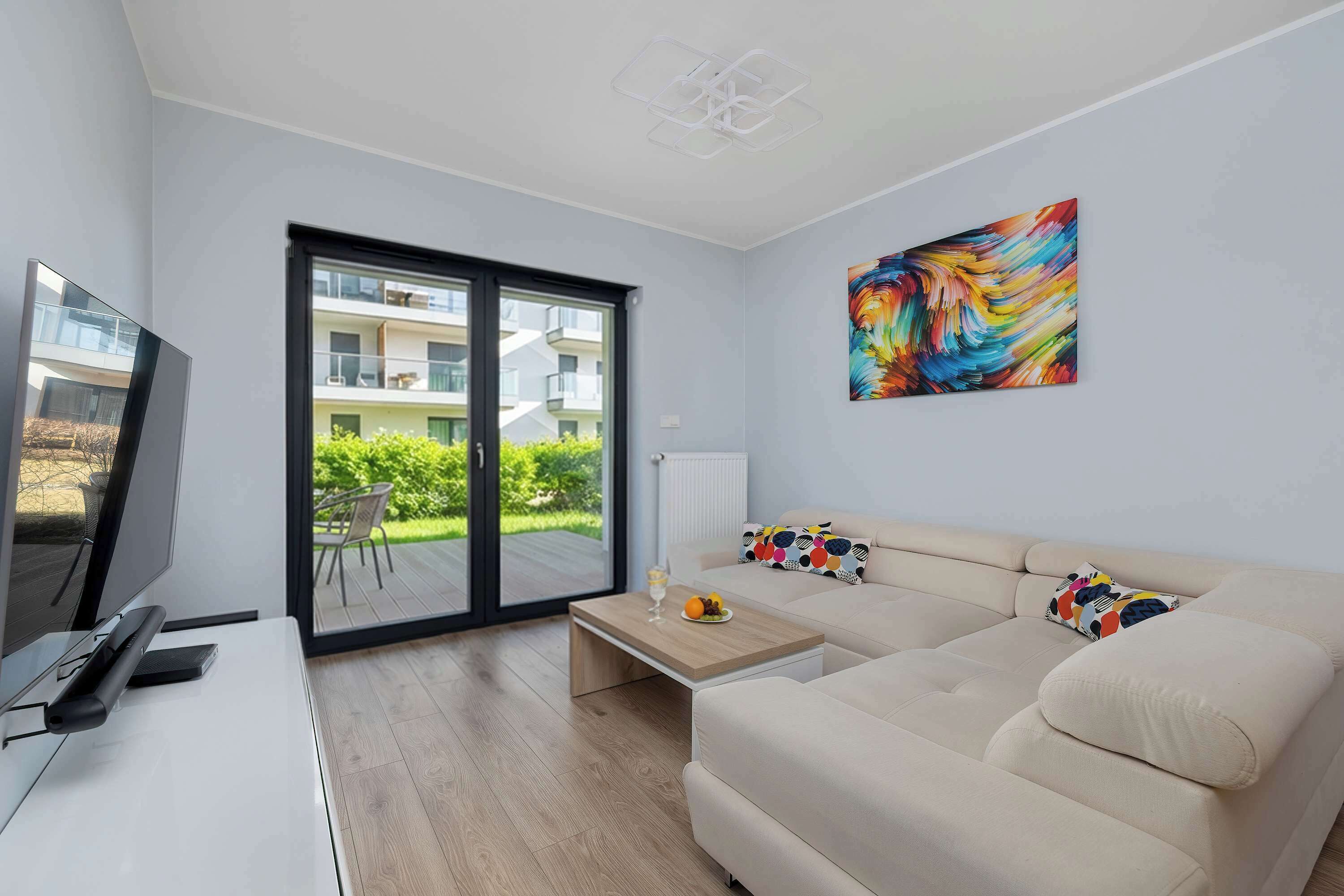 A spacious lounge view highlights the open-concept layout connecting the seating area to the garden. A large flat-screen TV is mounted on the wall across from the comfortable sectional sofa.
