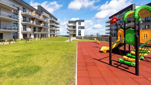 Un'area giochi con altalene e scivoli, circondata da moderni palazzi con balconi e una zona verde.