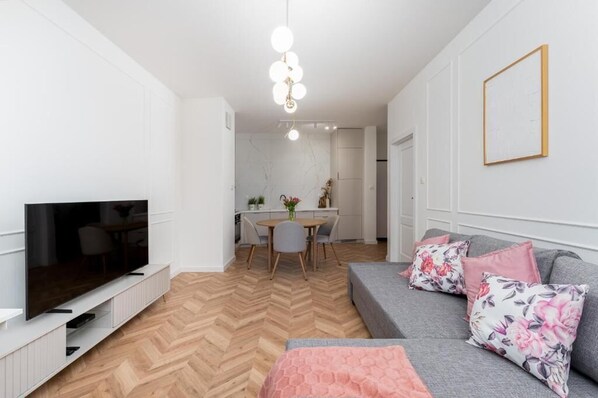 A modern living room with a flat-screen TV, a grey sofa with floral cushions, and a herringbone-patterned wooden floor. - Malila Apartments Cracow Rakowicka 254 (Kraków)