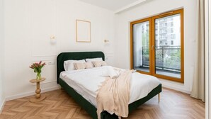 A bedroom with a bed, a small round table with a vase, and a window with a view of buildings. - Malila Apartments Cracow Rakowicka 254 (Kraków)