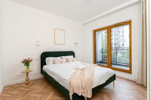 Ein Schlafzimmer mit einem Bett, einem kleinen runden Tisch mit einer Vase und einem Fenster mit Blick auf Gebäude.