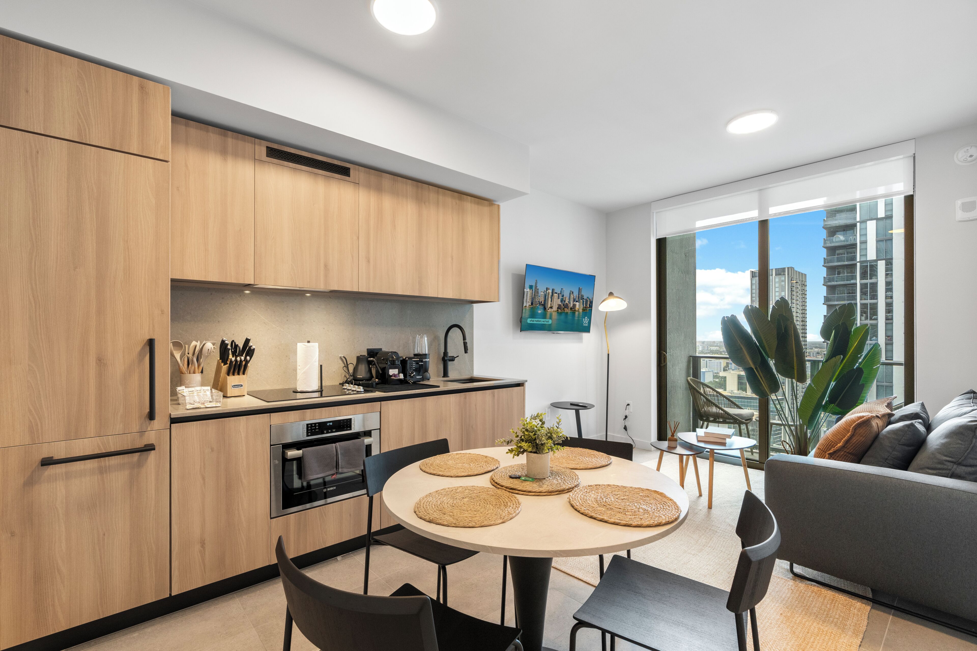 A modern kitchen with wooden cabinets, a dining area with a round table, and a living area with a sofa and large windows.