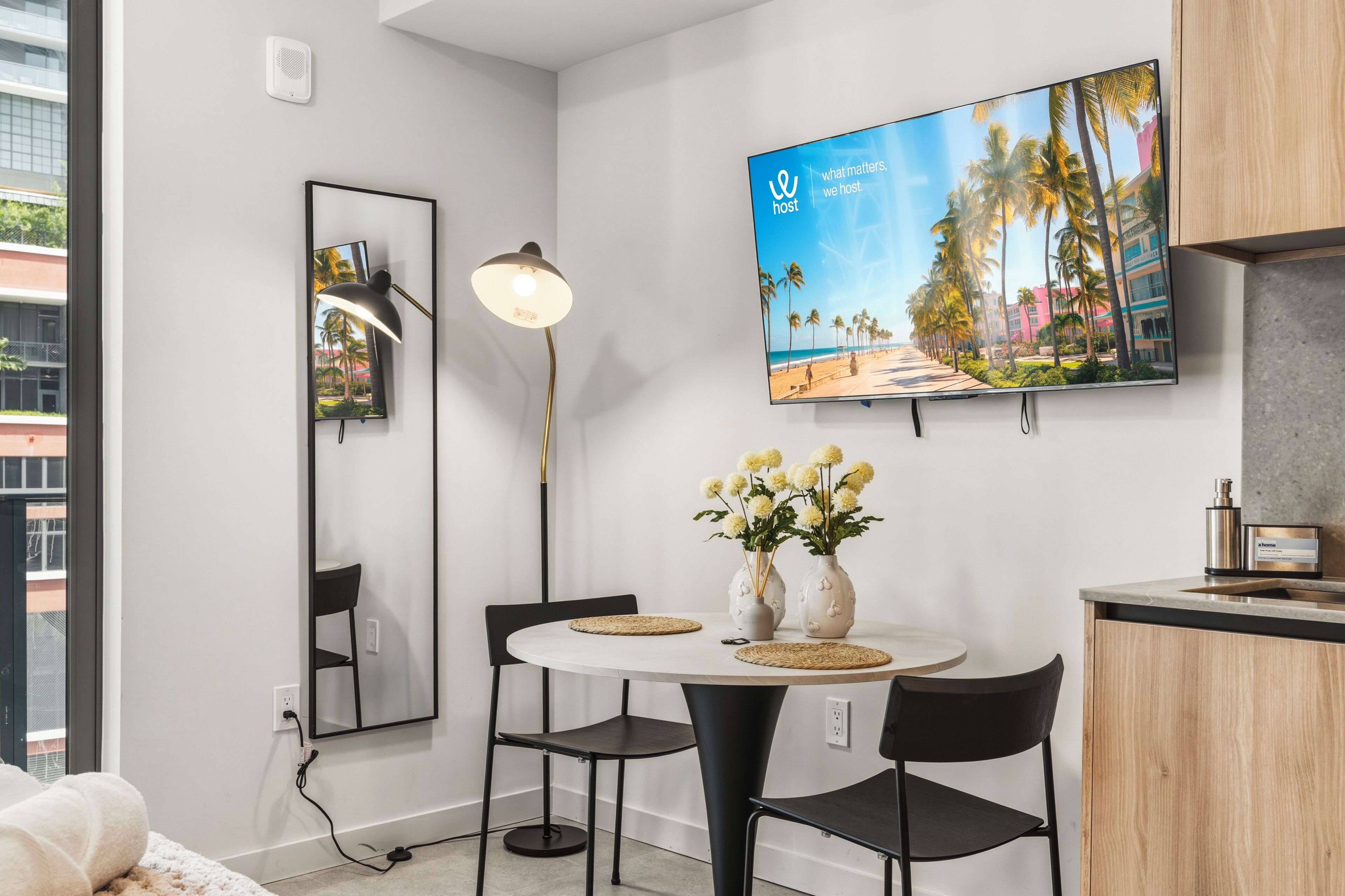 Une pièce moderne avec une table de salle à manger ronde, deux chaises, un téléviseur fixé au mur, une lampe sur pied et un petit coin cuisine.