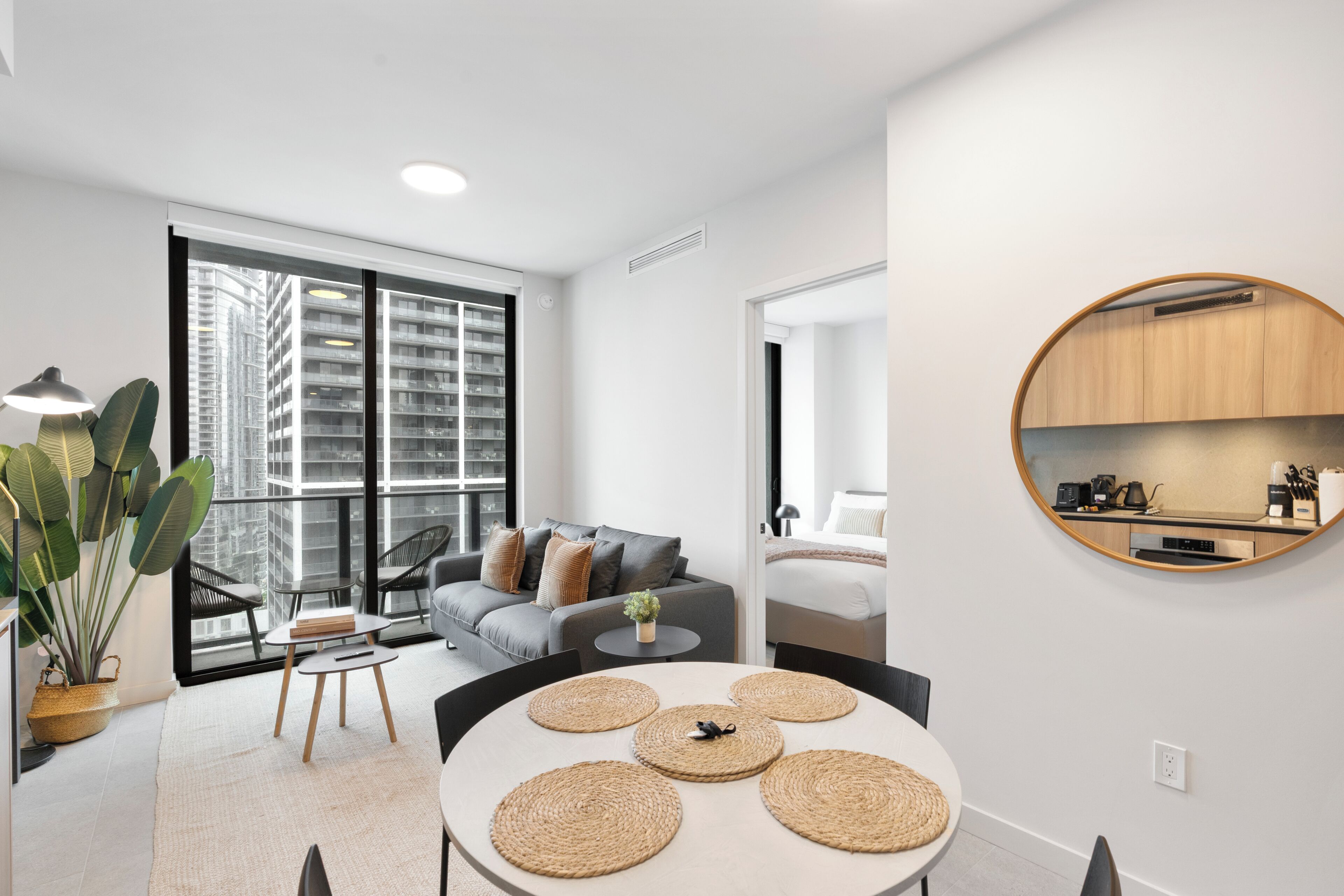 Ein modernes Wohnzimmer mit Sofa, Couchtisch und Esstisch, eine Küche mit rundem Spiegel und große Fenster mit Blick auf die Stadt.