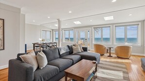Een ruime woonkamer met een grote grijze bank, een houten salontafel en uitzicht op de oceaan door grote ramen.