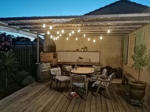 Eine Terrasse im Freien mit rundem Tisch und Stühlen, einem Backofen aus Ziegeln, Lichterketten und einem überdachten Sitzbereich.