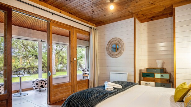 Et soveværelse med en stor seng, træloft, et vindue med udsigt til træer, og et lille bord med stol udenfor.
