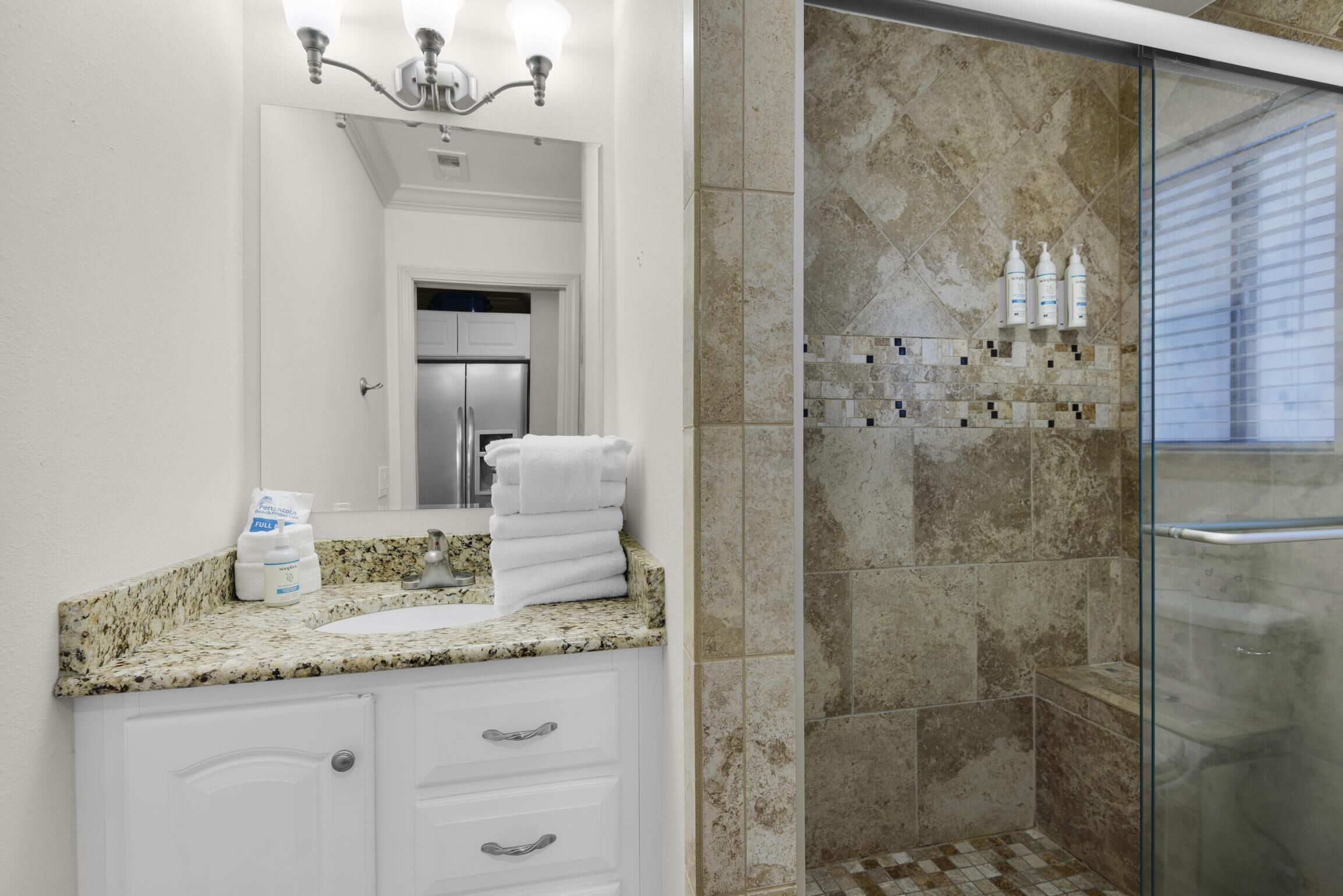 A bathroom with a granite countertop, white cabinets, a glass shower enclosure, and a tiled shower wall.
