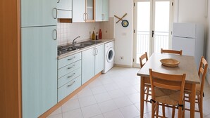 A compact kitchen with light blue cabinets, a sink, a stove, a washing machine, and a dining area with a table and chairs. - Appatamenti ABR Corso - Apartment with View and Balcony (Le Castella)