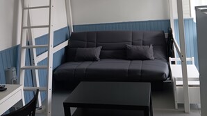 A bunk bed with a ladder, a sofa, a table, and a chair in a room with a window and a ceiling light fixture.