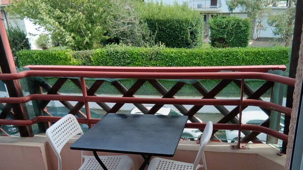 A balcony with a table and two chairs, surrounded by a red railing and a view of a residential area with houses and greenery.