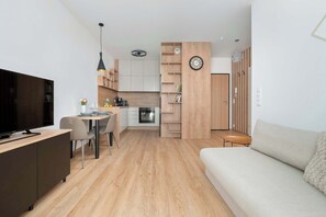 A modern living room with a sofa and a TV, a dining area with chairs and a table, and a kitchen with cabinets and a clock. - Bright Apartment in Warsaw | Private Garden and Parking (Warszawa)