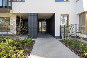 A modern building with a paved walkway, a vertical black panel, and a metal fence. - Bright Apartment in Warsaw | Private Garden and Parking (Warszawa)